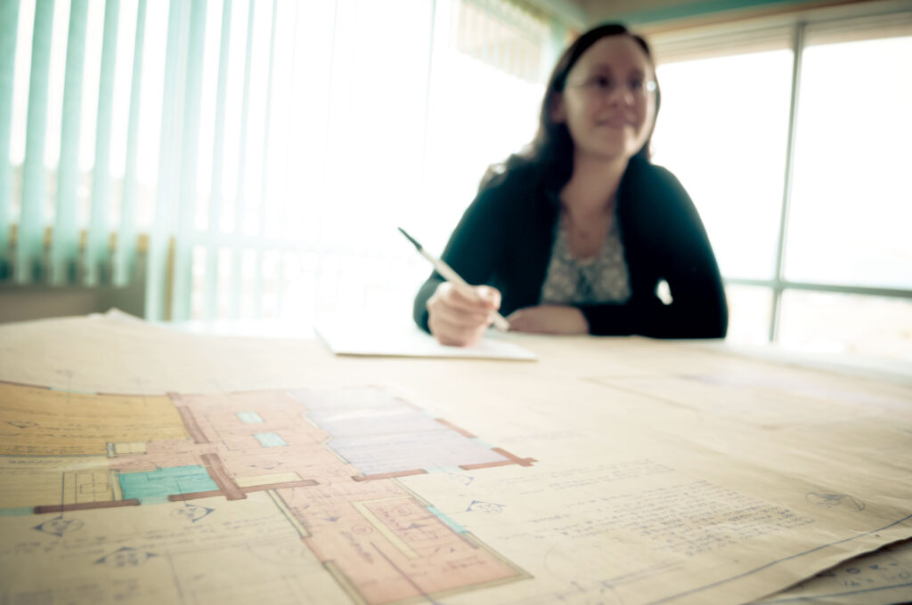 A woman in the background with a desk full of architectural plans lay in front of her