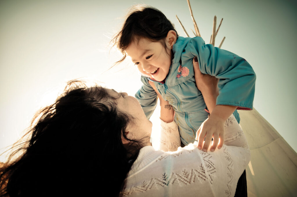 A woman holds up a child with a teepee in the background